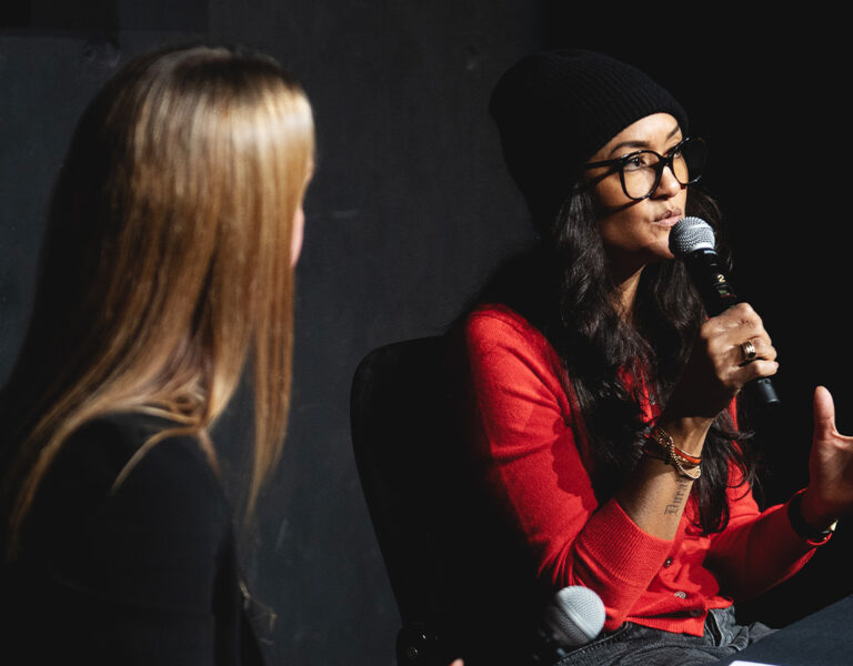 Two women sit at a table in conversation. One, wearing a red shirt, glasses, and a black beanie, gestures while speaking into a microphone during an event review. The other woman, with long blonde hair, faces her as they discuss Camerimage 2026.
