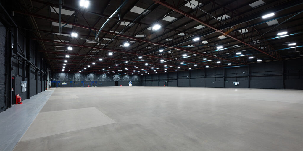 A large, empty indoor industrial space with high ceilings, exposed metal beams, and evenly spaced overhead lights illuminating the concrete floor—ideal for studios or industry wide shot set-ups. Dark walls feature several fire extinguishers mounted along the sides.