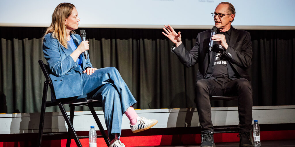 At the Mallorca International Film Festival, a woman in a blue suit and a man in black clothing sit on stage, each holding a microphone. Engaged in conversation, they have water bottles on the floor beside their chairs.