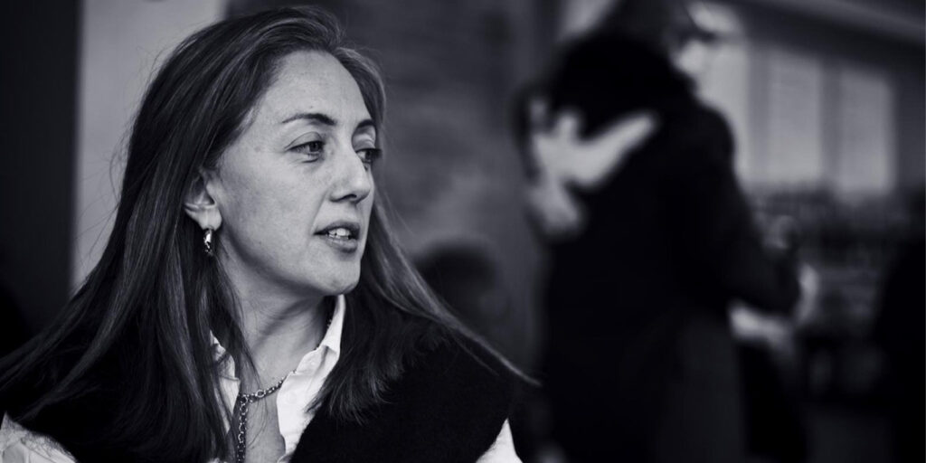 A woman with long hair and a necklace looks to her left in the foreground. The background is blurred, with two people hugging near a window. This black and white image captures a moment of evolution in the world of CineVoices.