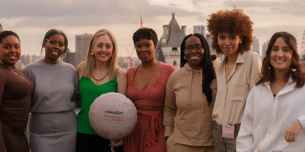 Seven women stand together on a rooftop at sunset, smiling at the camera. One holds a round balloon printed with words. City buildings and a cloudy sky create the backdrop for this moment of Cinegirl evolution.
