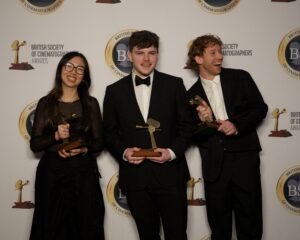 Three people in formal wear stand in front of a British Society of Cinematographers Awards backdrop, each holding a trophy and smiling for the camera.