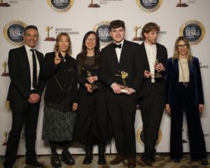 Six people stand in a row at an awards event in front of a British Society of Cinematographers backdrop. Three people in the middle are holding trophies. Everyone is dressed in formal or semi-formal attire.