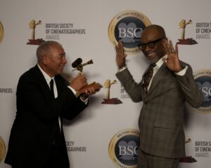Two men in suits pose in front of a British Society of Cinematographers Awards backdrop; one holds a trophy and points it playfully at the other, who smiles and raises his hands in a cheerful gesture.