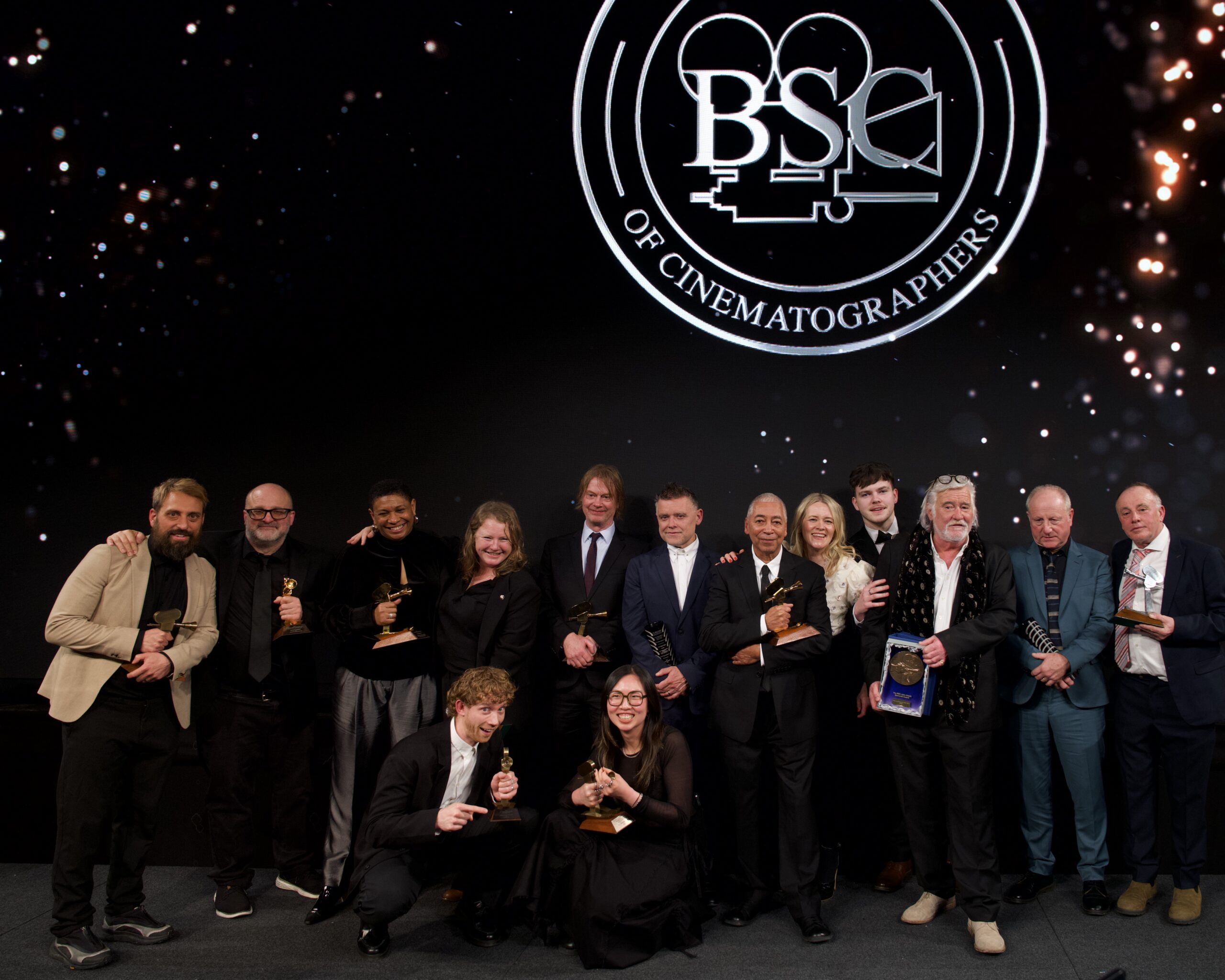 A group of people pose on stage holding awards in front of a large screen displaying the British Society of Cinematographers logo. Some are standing, whilst others are kneeling or crouching.