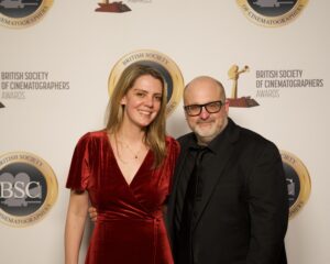A woman in a red velvet dress stands next to a man in a black suit and glasses, both smiling, in front of a British Society of Cinematographers Awards backdrop.