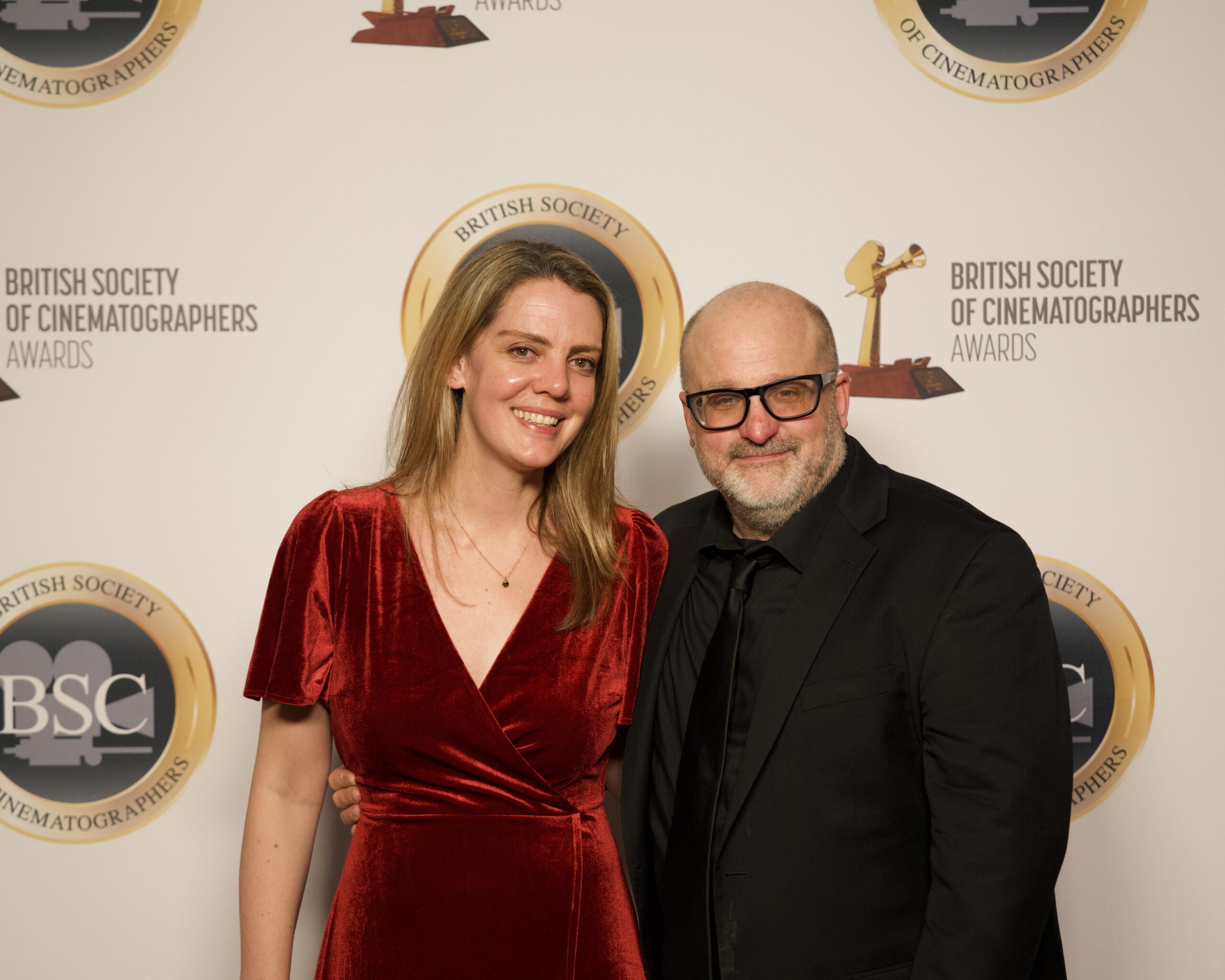 A woman in a red velvet dress stands next to a man in a black suit and glasses, both smiling, in front of a British Society of Cinematographers Awards backdrop.