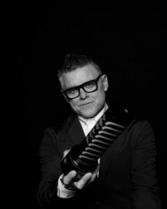A person with short, styled hair and glasses, dressed in a dark suit and white shirt, holds a large cylindrical object towards the camera against a black background.