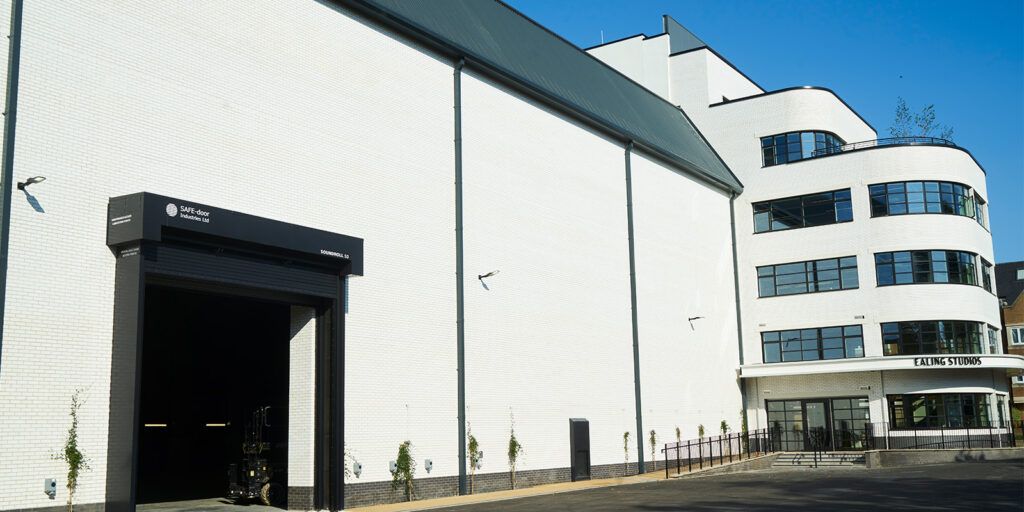 A large white building with modern, curved architecture and black trim, labelled Ealing Studios above the main entrance, is shown in an industry-wide shot under a clear blue sky. A loading bay with an open door is visible on the left.