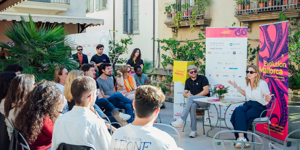 A woman and a man speak to a seated audience in an outdoor courtyard at the Mallorca International Film Festival, surrounded by banners and potted plants. The audience listen attentively, with a few standing at the back.