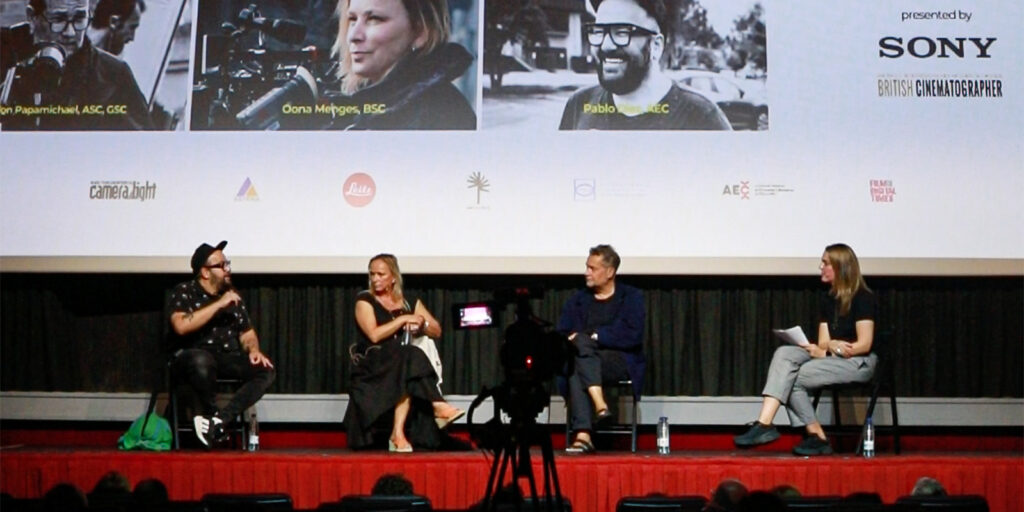 Four people sit on stage in front of a screen during a panel discussion at the Evolution Mallorca Film Festival. One gestures whilst speaking, others listen intently. Audience heads and a centrally placed camera are visible in the foreground.