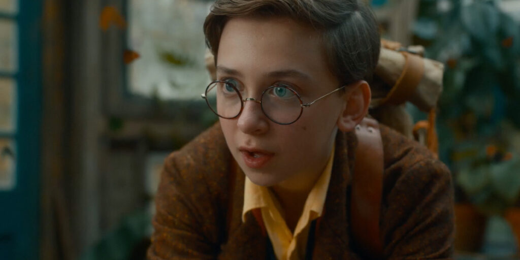 A young person with short brown hair, round glasses, and a brown jacket looks ahead with a focused expression. Reminiscent of Caroline Bridges from The Famous Five, they wear a yellow shirt and have a rolled-up item strapped to their back. Green plants are in the background.