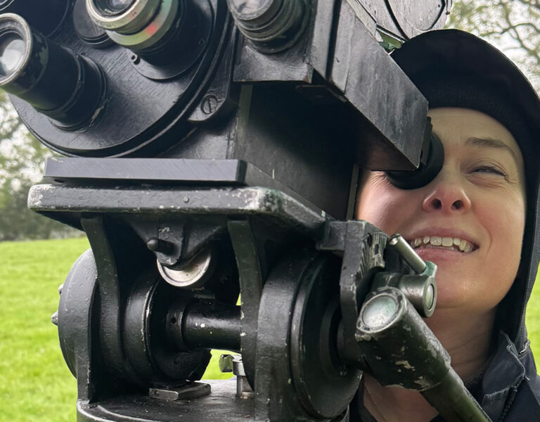 A person in outdoor clothing, reminiscent of Caroline Bridges from The Famous Five adventures, looks through the eyepiece of a large black vintage camera on a grassy field, with trees visible in the background.