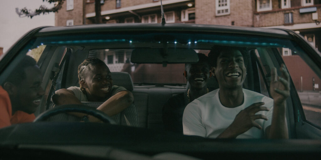 Four young people sit and smile inside a car, two in the front seats and two in the back, talking and laughing. A block of flats and street are visible through the windscreen in this Film Soho sponsored feature.