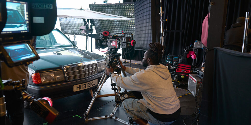 A person adjusts a camera on a tripod in front of a green car with the number plate H113 EKJ inside Film Soho's studio, where lighting equipment and tools are visible around the set for this sponsored feature.