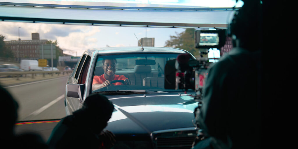 A man sits in the driver’s seat of a car, smiling and holding the steering wheel, as a Film Soho crew with camera equipment captures this sponsored feature on a bustling carriageway.