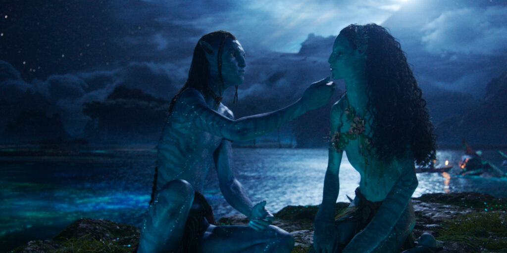 Two blue-skinned humanoids from Avatar: Fire and Ash sit by a glowing, moonlit lake. One gently touches the other’s chin as Russell Carpenter’s cinematography brings alive the luminous water, distant trees, and cloudy night sky.