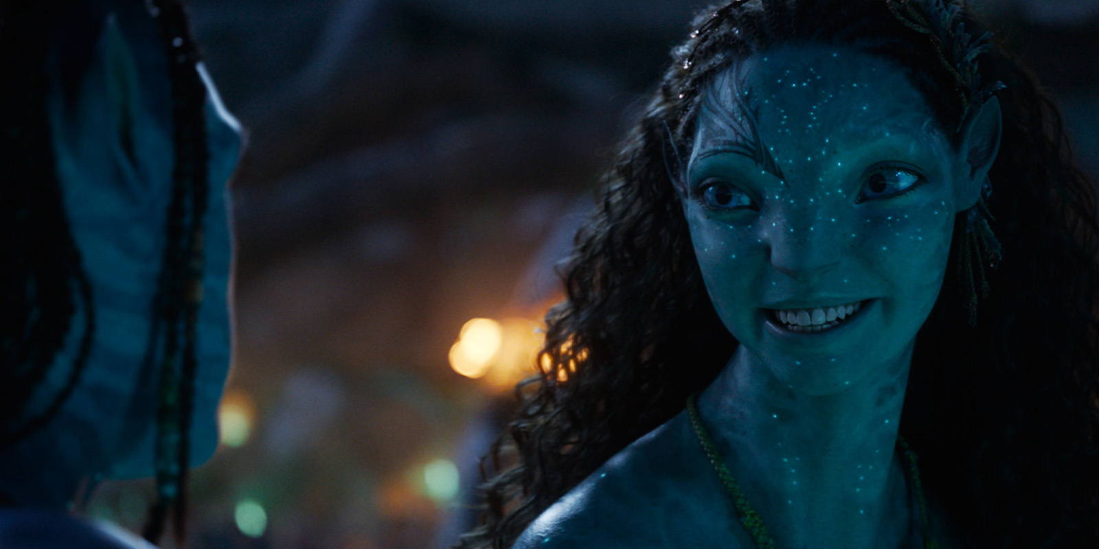 A blue-skinned, humanoid character with bioluminescent spots, yellow eyes, and curly hair smiles whilst facing another similar figure in a scene reminiscent of Russell Carpenter’s work on Avatar: Fire and Ash, softly lit with warm, out-of-focus lights.