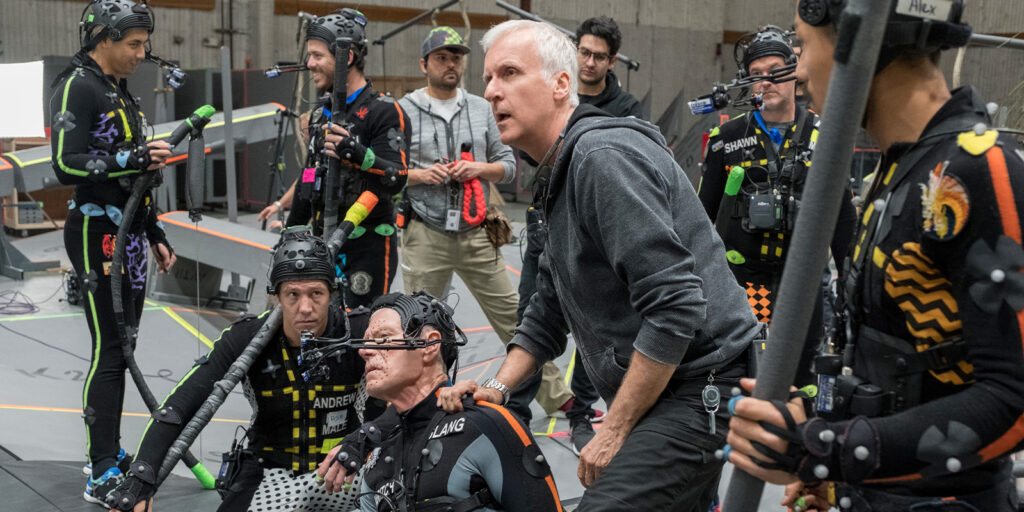 A group of people on a motion capture set—some in sensor suits and headgear—while a man in a grey hoodie, possibly directing the group during the production of Avatar: Fire and Ash, stands among them.