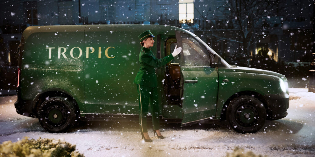 A person in a green uniform stands beside a green van labelled TROPIC, open door spilling light onto the snowy ground at night. Snow covers garden studios and the surrounding area, blending softly into the wintry scene.