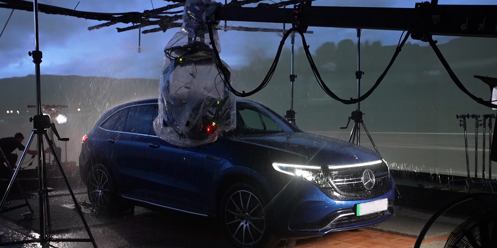 A blue Mercedes-Benz SUV is parked under rigged equipment and plastic-wrapped cameras at Garden Studios, as artificial rain falls, suggesting a controlled environment for filming or testing in wet conditions.