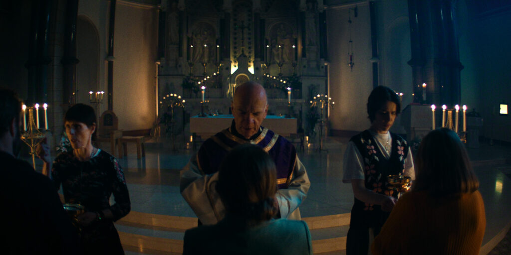 A priest stands at the altar of a church, accompanied by two people holding communion vessels, as he prepares to serve communion to a group of congregants facing him. The illuminated altar evokes peace, reminiscent of Ashley Barron's "How to Get to Heaven From Belfast.