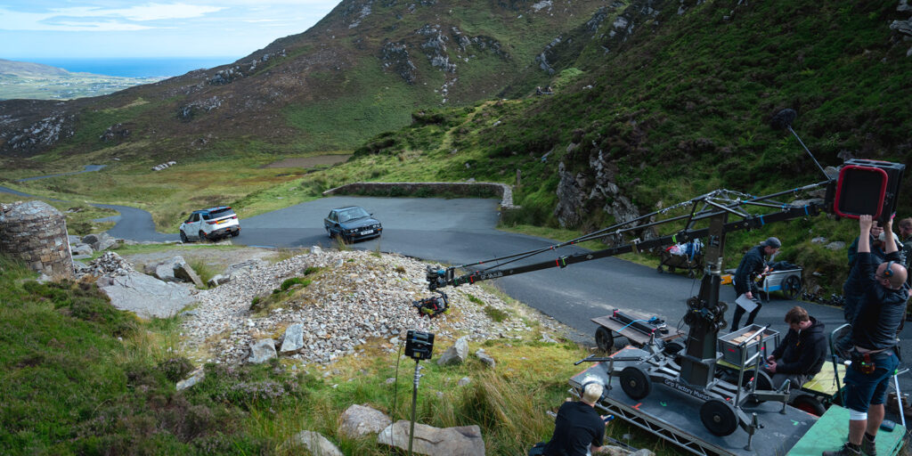 A film crew operates camera equipment on a mountain road, filming scenes for How to Get to Heaven From Belfast. Two cars sit on the winding carriageway as ACS crew members work among rocky terrain and grassy slopes.