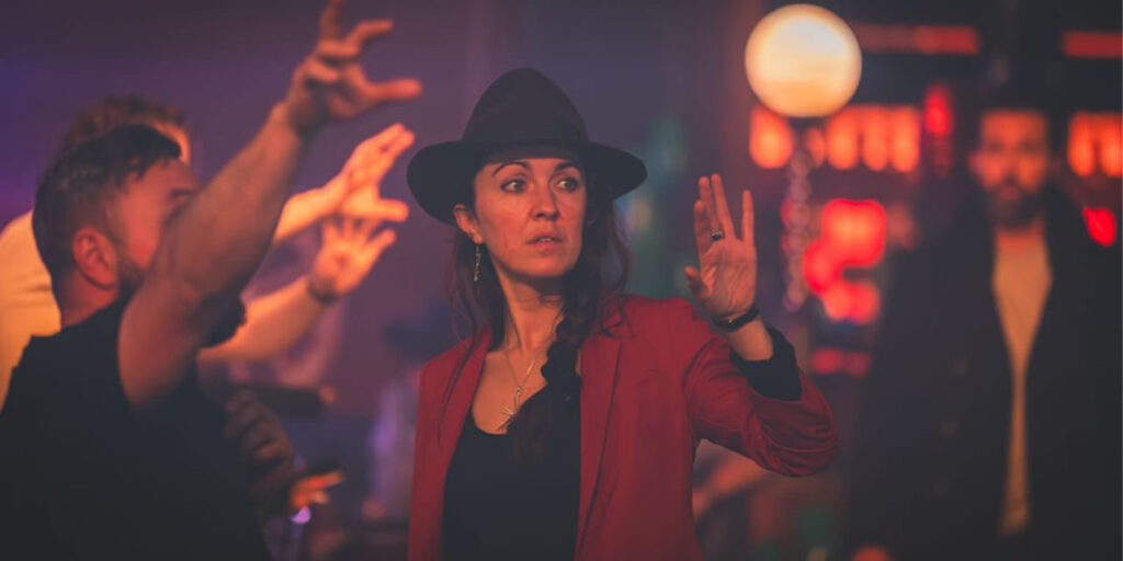 A woman in a black hat and red jacket stands indoors, raising her hand with a serious expression—like a scene from "How to Get to Heaven From Belfast." Around her, people gesture as dim, blurred lights fill the background.