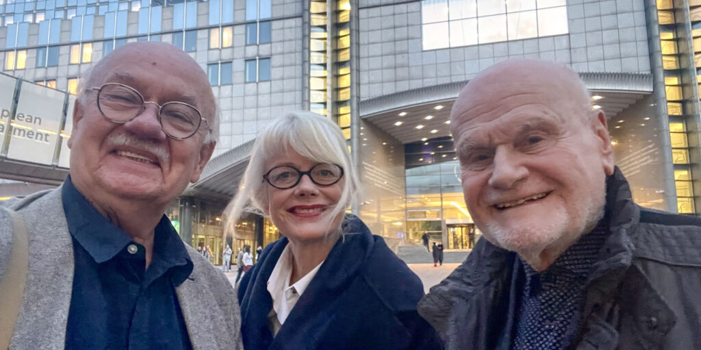 Three older adults, two men and one woman, are smiling and posing for a selfie outside a modern glass building with people in the background, capturing a joyful moment Behind the Lens for an IMAGO study.