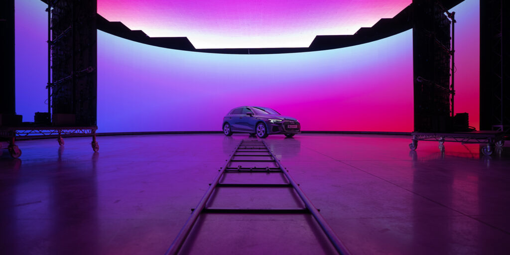 A car is parked in front of a large curved LED screen displaying a gradient of purple, pink, and blue colours. An industry wide shot captures the camera track on the floor leading towards the car in this vibrant indoor studio setting.