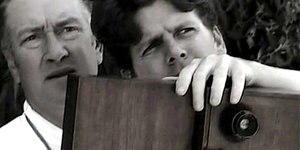 Two people stand close together outdoors, intently looking in the same direction. The person in front, reminiscent of a scene from Mulholland Drive, holds a large, boxy wooden camera that partially obscures their face. The photo is in black and white.