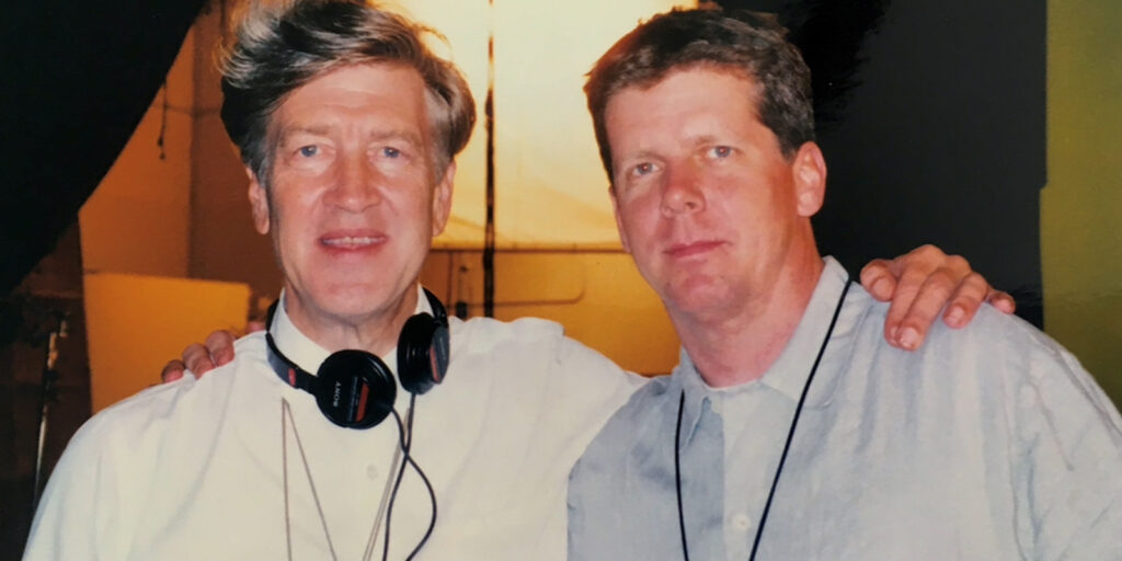 Two men stand side by side, both wearing light-coloured shirts. One man, with headphones around his neck and his arm around the other’s shoulder, evokes a behind-the-scenes moment on a Peter Deming set. They face the camera against a bright background.