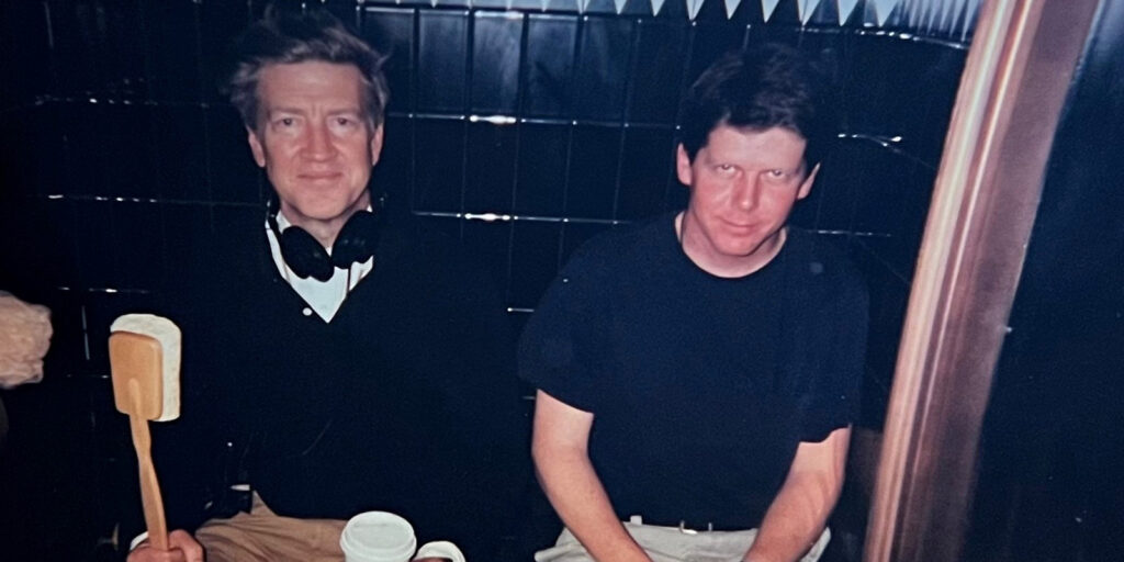 Two men are sitting side by side in a dark, tiled setting reminiscent of Mulholland Drive. The man on the left, possibly Peter Deming, holds a wooden spatula and cup, with headphones round his neck. Both are dressed casually.