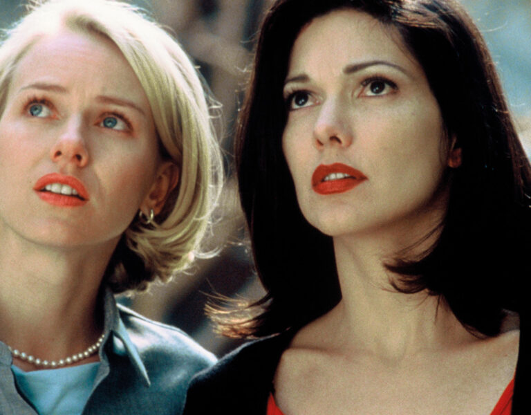 Two women with serious expressions stand close together outdoors, looking slightly upward—a scene reminiscent of Peter Deming’s cinematography in Mulholland Drive. One has short blonde hair and pearls; the other wears a black top, red lipstick, and has shoulder-length dark hair.