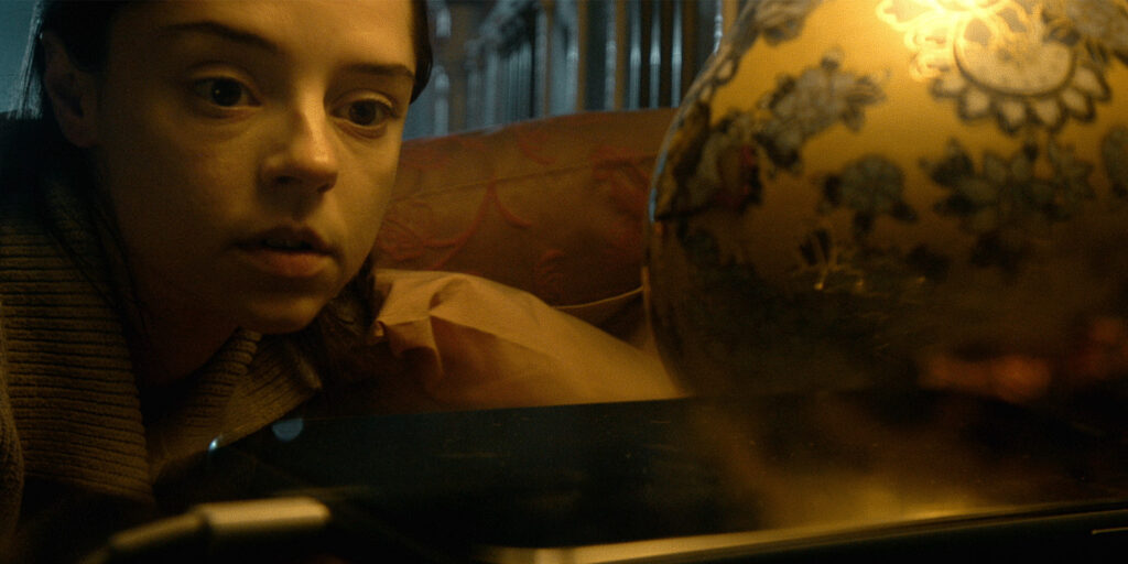 A person with a serious expression looks closely at a shiny, reflective surface beside a floral-patterned lamp in a dimly lit room, as Outside Noise seems to echo softly in the warm, yellowish glow.