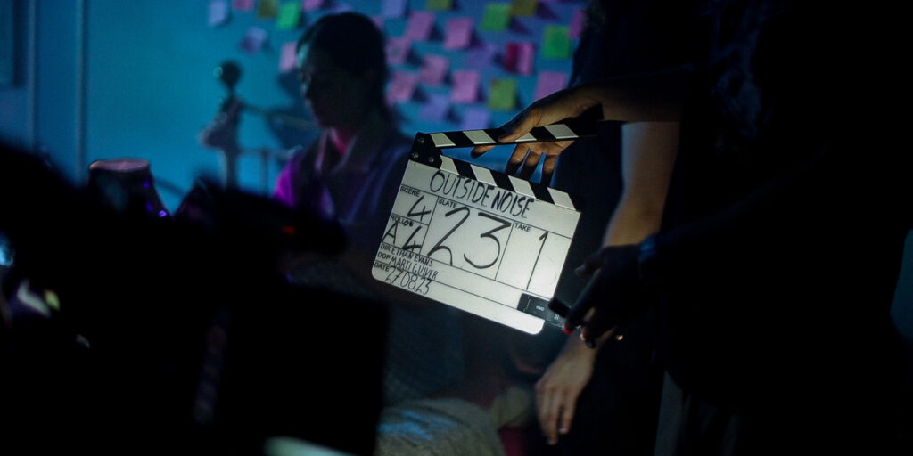 A person holds a clapperboard labelled Outside Noise in front of a dimly lit set; Marti Guiver is a blurred figure in the background, and colourful sticky notes line the wall.