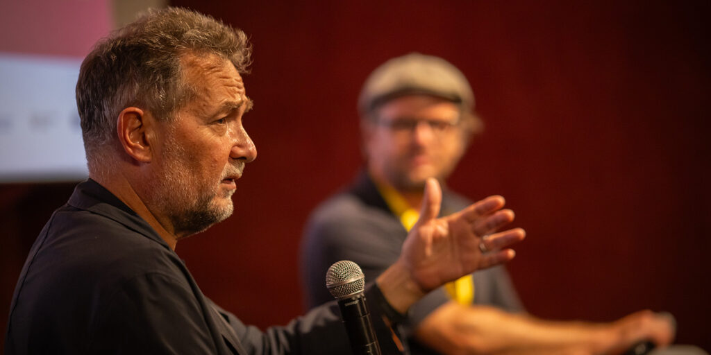 A man in focus speaks into a microphone while gesturing with his hand at the Mallorca International Film Festival; another man, blurred in the red-toned background, listens. Both are indoors.