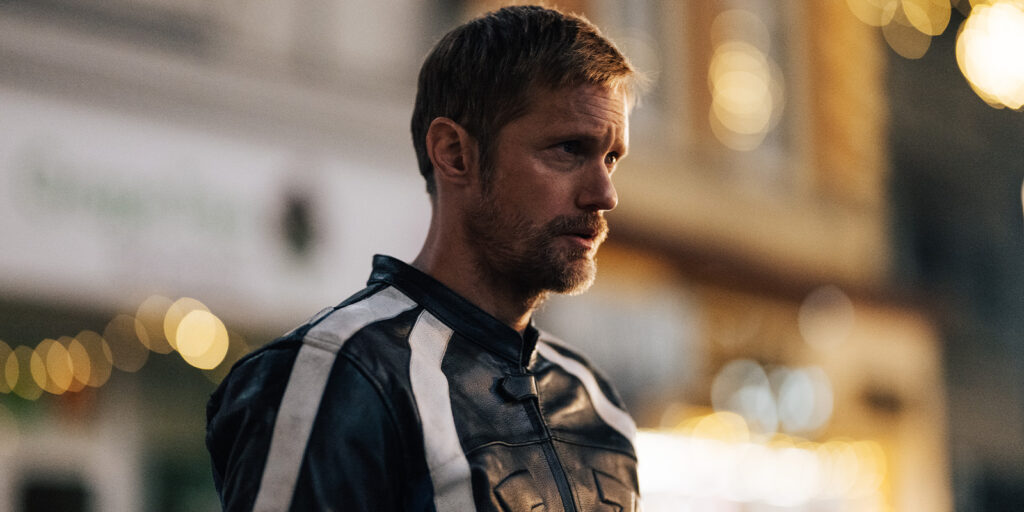 Nick Morris, a man with short hair and a beard, stands outdoors wearing a black jacket with white stripes. The blurred background features glowing lights and buildings, creating a vibrant scene for this Pillion moment.