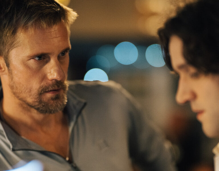 A man with short hair and a beard, possibly Nick Morris, looks intently at another man with dark, wavy hair, who is slightly out of focus. The blurred lights in the background suggest an indoor pillion setting.