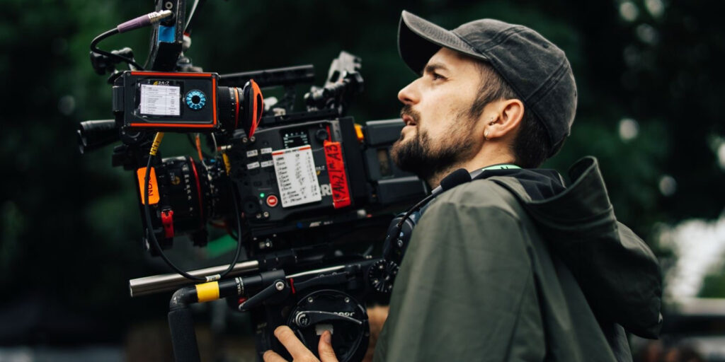 A person wearing a cap and jacket operates a professional video camera outdoors, focusing intently as they film. The camera, equipped with various attachments and a monitor, captures the scene as trees blur behind—possibly on set with Nick Morris.