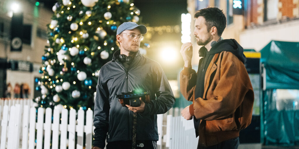Two men, including Nick Morris, stand outside at night near a decorated Christmas tree and a white fence, talking. One holds a device with headphones around his neck. The festive lights and buildings create a warm pillar of holiday cheer in the background.