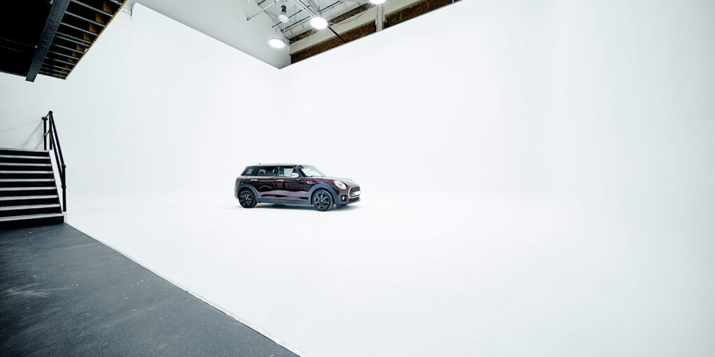 A black compact car is parked in the centre of a large, white, empty studio space with high ceilings and overhead lights—a true industry wide shot. A staircase is visible on the left side of the image.