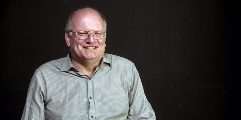 A middle-aged man with glasses, short fair hair, and a light grey button-up shirt smiles whilst sitting in front of a plain dark background, ready to present his annual summary.