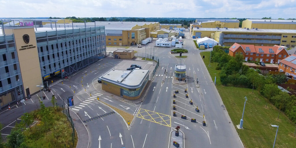 Aerial industry wide shot of a large industrial complex with multiple yellow buildings, studios, a multi-storey car park, a guardhouse, marked roads, and adjacent red-roofed houses on a partly cloudy day.