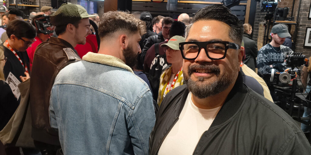 A man with glasses, a beard, and a black jacket smiles at the camera during the busy BSC Expo 2026. Other people in casual clothes, some in hats and jackets, mingle nearby as camera equipment fills the background.
