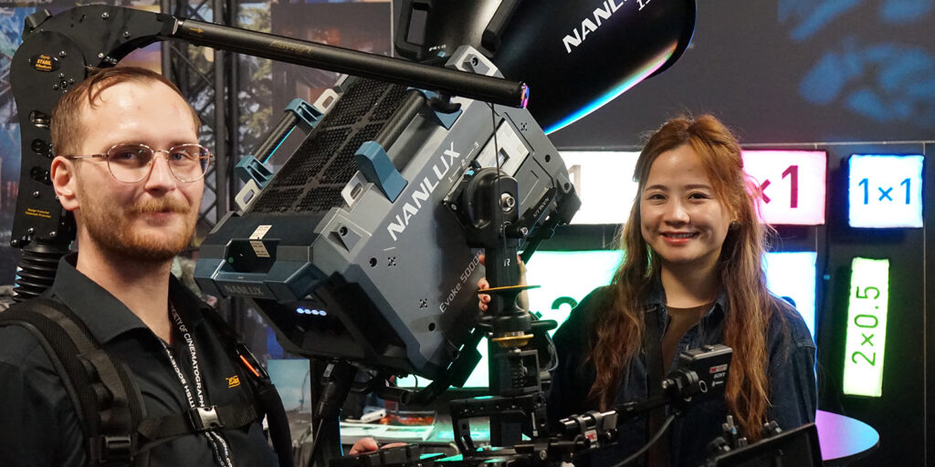 At the BSC Expo 2026, a man wearing glasses and a black shirt stands next to a large NANLUX film light. Beside him, a smiling woman with long hair poses as colourful "1x1" and "2x0.5" signs glow in the background.