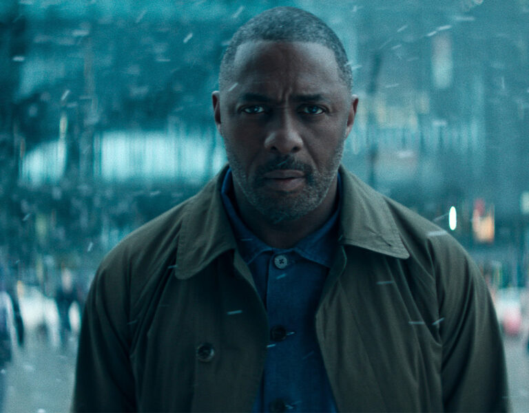 A man with a serious expression stands outdoors in a busy, snow-filled urban setting. He wears a brown coat over a blue shirt whilst people are blurred in the background.