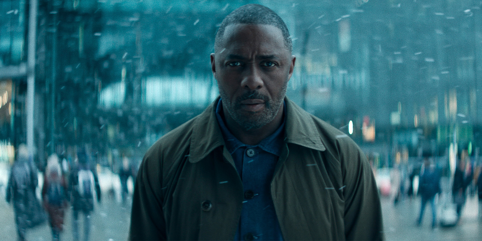 A man with a serious expression stands outdoors in a busy, snow-filled urban setting. He wears a brown coat over a blue shirt whilst people are blurred in the background.