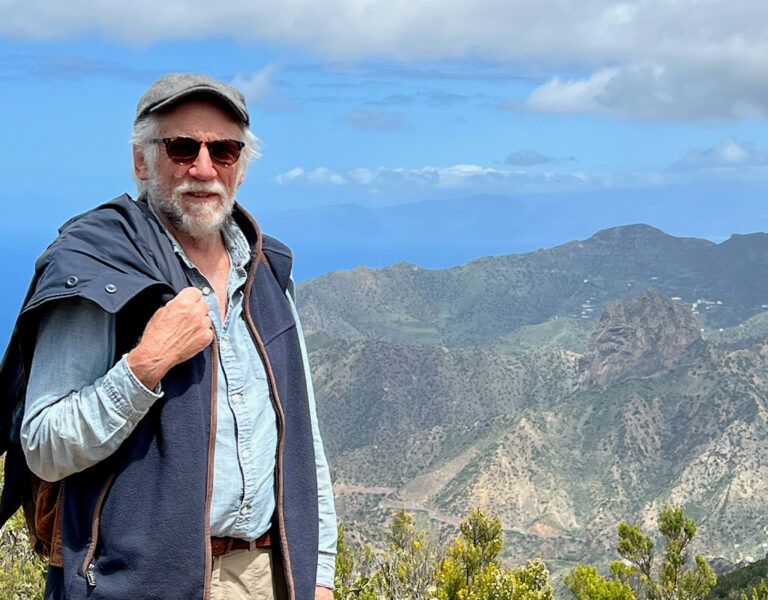 An older man wearing a cap, sunglasses, and a jacket draped over his shoulder stands on a mountain path with a view of green hills, cliffs, and distant sea under a partly cloudy sky.