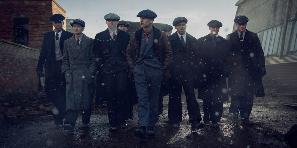A group of nine men in early 20th-century attire, including flat caps and long coats, walk together on a wet, debris-strewn street in an industrial setting, with light snow or rain falling.
